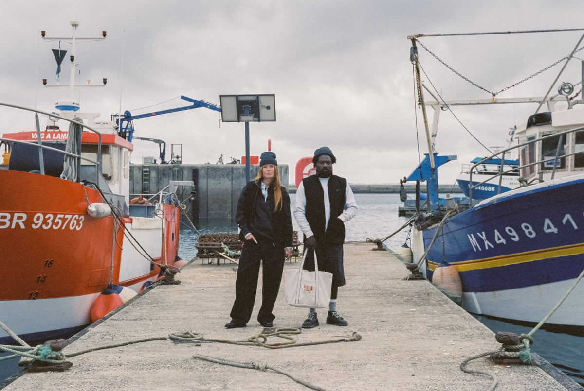 Deux personnes en bonnets Youtar 1894 sur un ponton du port de Brest, entre deux chalutiers, avec un tote bag Youtar